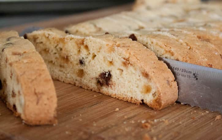 A loaf of freshly baked Toffee Chocolate Chip Biscotti being sliced on a cutting board by themerchatbaker.com