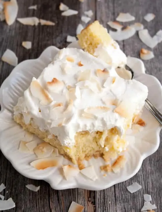A piece of Pineapple Coconut Tres Leches Cake on a fork resting on serving dish with coconut sprinkled around the serving dish from themerchantbaker.com