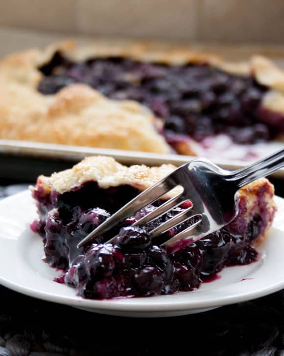 A slice of Blueberry Galette on a pate with a fork taking a piece from themerchantbaker.com