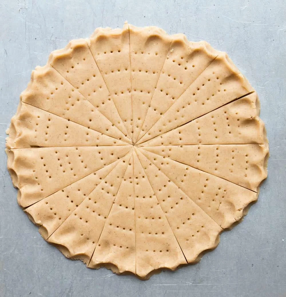Raw dough in a circled shape with pinched edges, pressed with a fork and cut crosswise on a baking sheet to make Spiced Shortbread by themerchantbaker.com