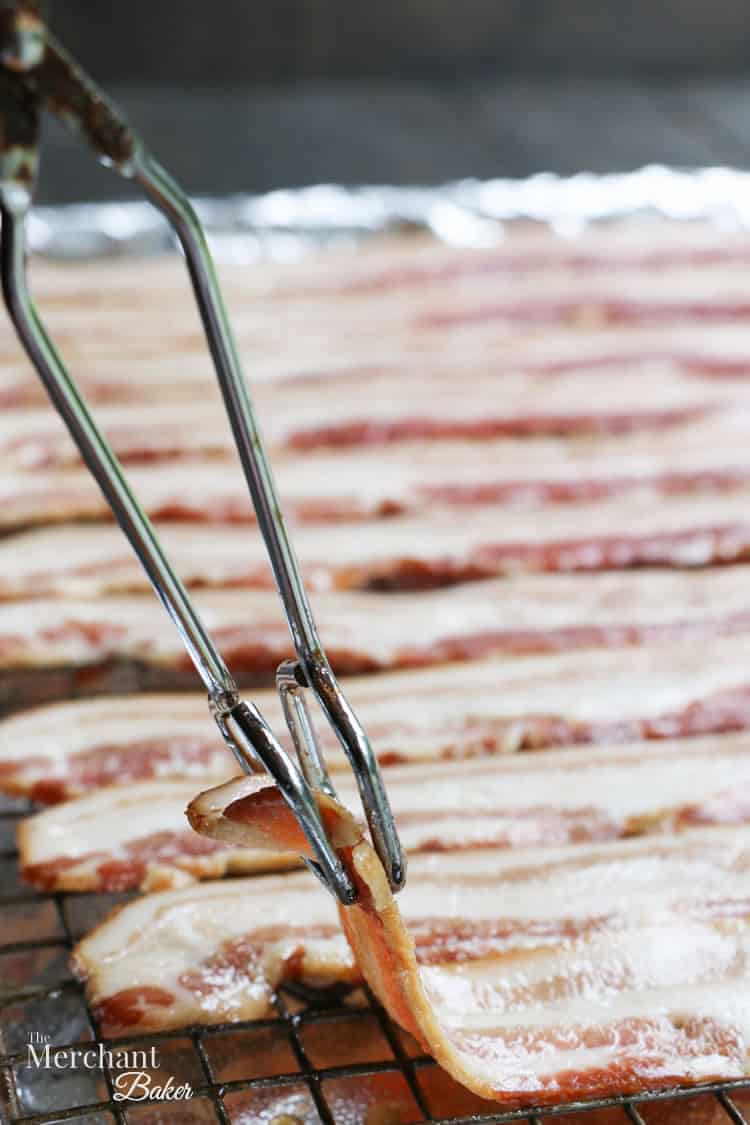 Lifting raw bacon with tongs to show thickness for Brown Sugar Maple Glazed Bacon from themerchantbaker.com