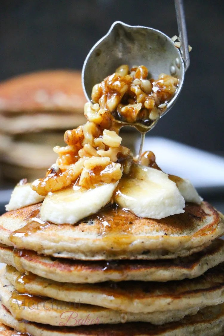 Walnut syrup being ladled over a stack of Oatmeal Chocolate Chip Banana Pancakes topped with sliced bananas 