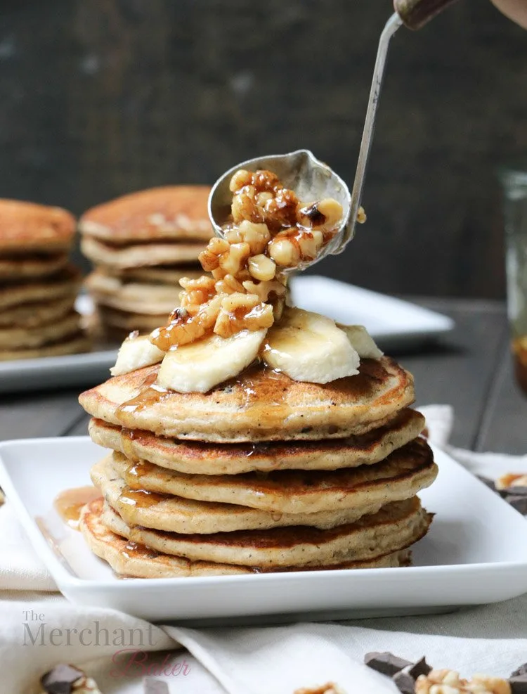 Walnut syrup being ladled over a stack of Oatmeal Chocolate Chip Banana Pancakes topped with sliced bananas