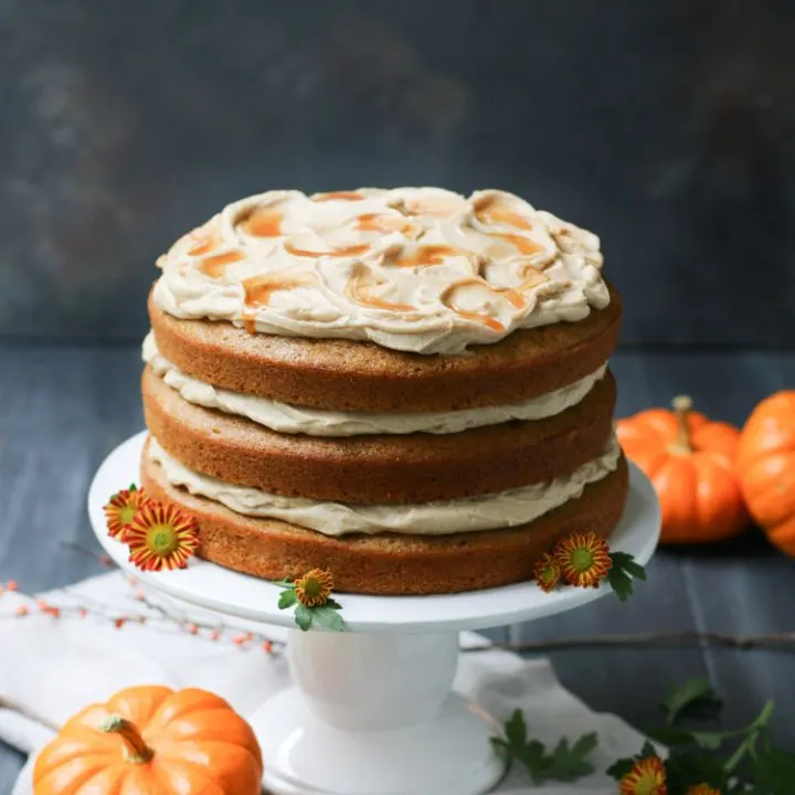 A high angle photo of a three tiered Overhead photo of caramel within icing on a Pumpkin Ale with icing on a pedestal with small pumpkins Cake by themerchantbaker.com