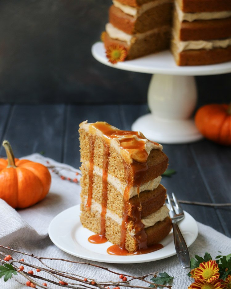 A slice of Pumpkin Ale Cake with caramel sauce on a small plate with Pumpkin Ale Cake on a pedestal in the background by themerhantbaker.com