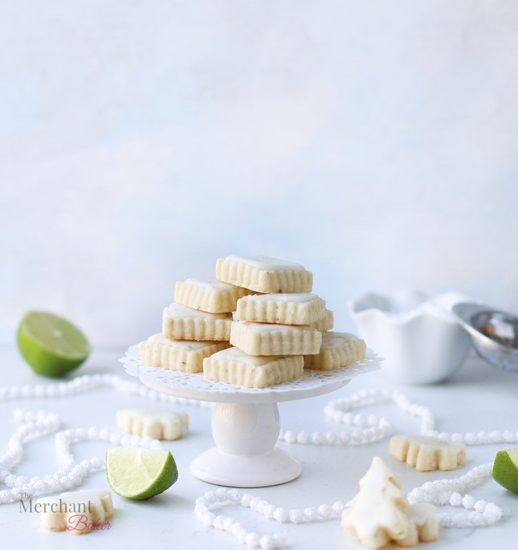 A stack of lime meltaway cookies on a small cake stand surrounded by tree shaped cookies and pieces of cut up lime