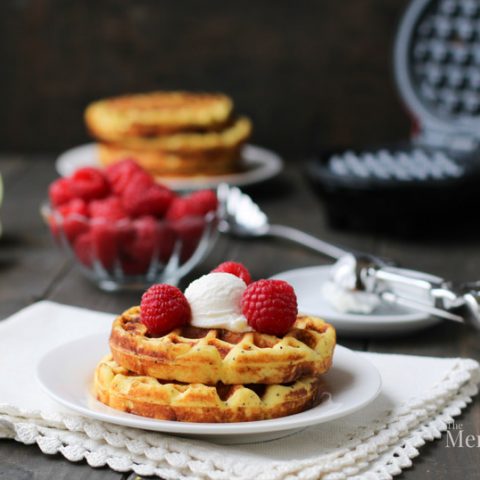 Side view of Lemon Ricotta Poppyseed Chaffles on a white plate with raspberries and a scoop of ricotta cheese on top by themerchantbaker.com