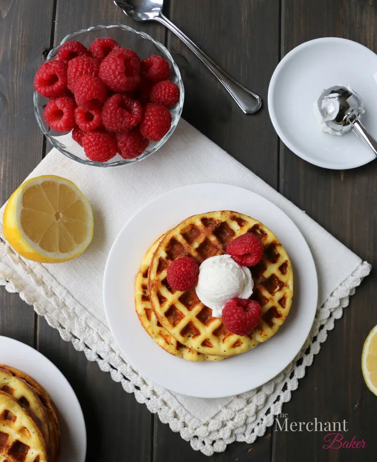 Overhead view of Lemon Ricotta Poppyseed Chaffles topped with raspberries and a scoop of ricotta with a bowl of raspberries and half of lemon by themerchatbaker.com