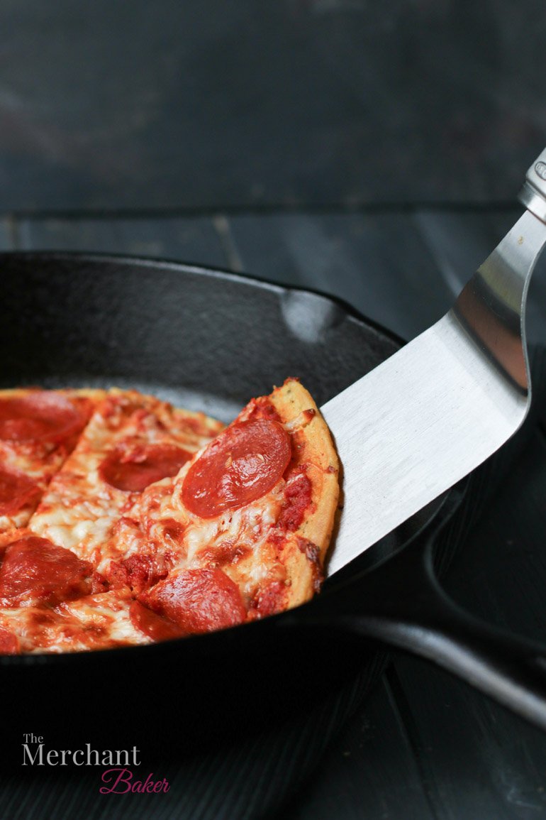 A side view of a spatula lifting a slice of Socca Pizza out of an iron skillet by themerchantbaker.com