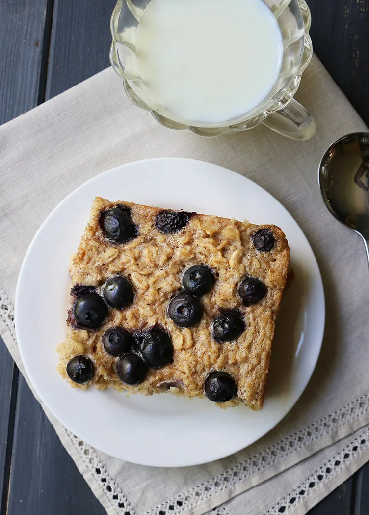 Overhead view of a square of baked oatmeal with blueberries on top next to a pitcher of milk 