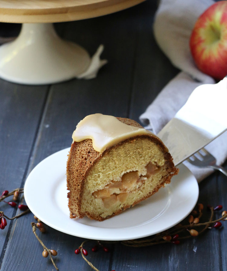 A slice of apple pie bundt cake being served onto a white plate by themerchantbaker.com