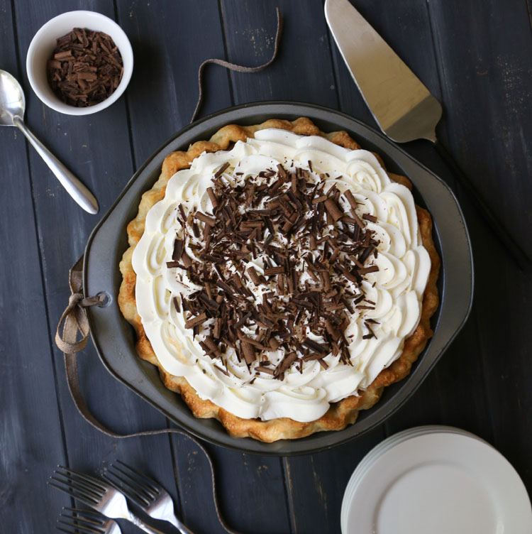Overhead photo of whole chocolate cream pie with plates, forks, pie server and bowl of chocolate shavings