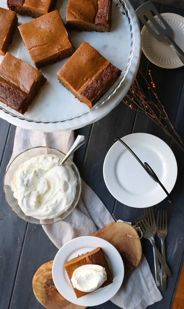 overhead view of pumpkin pie gingerbread cake on a white plate with a dollop of whipped cream on top sitting in front of a bowl of whipped cream and serving plates