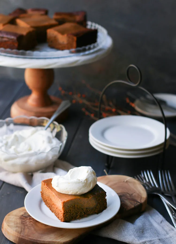 pumpkin pie gingerbread cake on a white plate with a dollop of whipped cream on top sitting in front of a bowl of whipped cream and serving plates