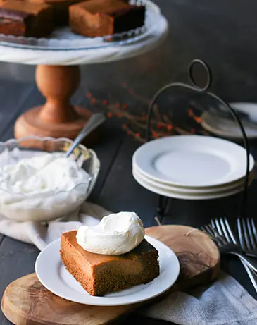 pumpkin pie gaingerbread cake on a white plate with a dollop of whipped cream on top sitting in front of a bowl of whipped cream and serving plates