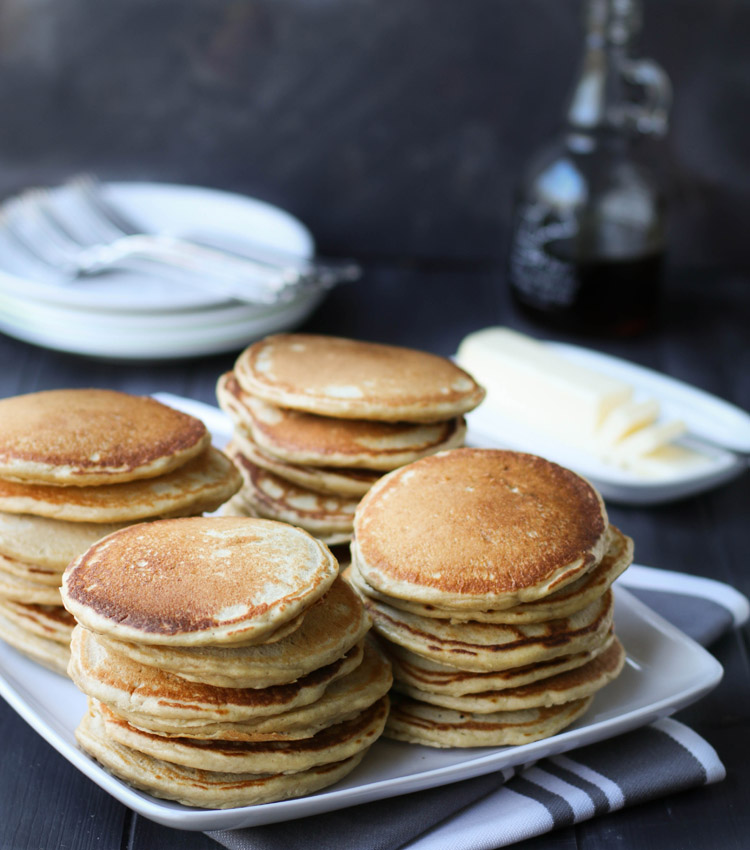 Four stack of pancakes on a serving platter with butter and syrup in the background