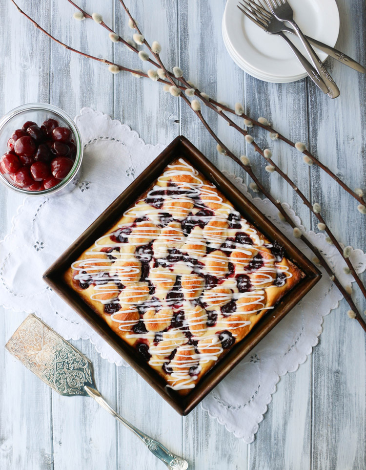 Overhead view of Cherry Cheese Danish Cake uncut in a square baking pan with container of cherries and a spatula beside it