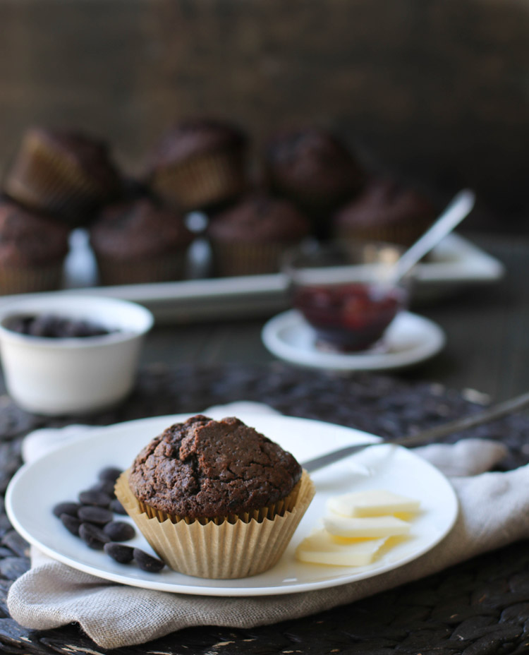 Chocolate muffin sitting on a white plate with pats of butter and dark chocolate chips. There is a tray of muffins and a dish of strawberry jam in the back ground.