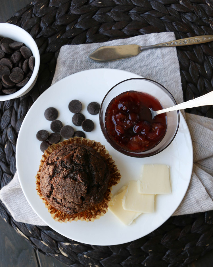 Overhead view of a white plate with a chocolate muffin, dark chocolate chips, strawberry jam and butter on it