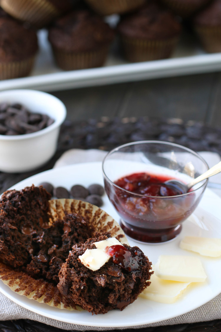 Chocolate muffin pulled open into halves spread with butter and jam sitting on a white plate with pats of butter and dark chocolate chips and a dish of jam on the plate.