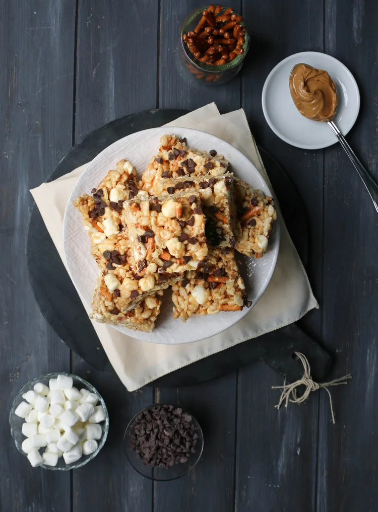 overhead view of peanut butter pretzel rice krispie treat bars stacked on a white plate surrounded by containers of pretzel stick, mini marshmallows, mini chocolate chips and a spoonful of creamy peanut butter