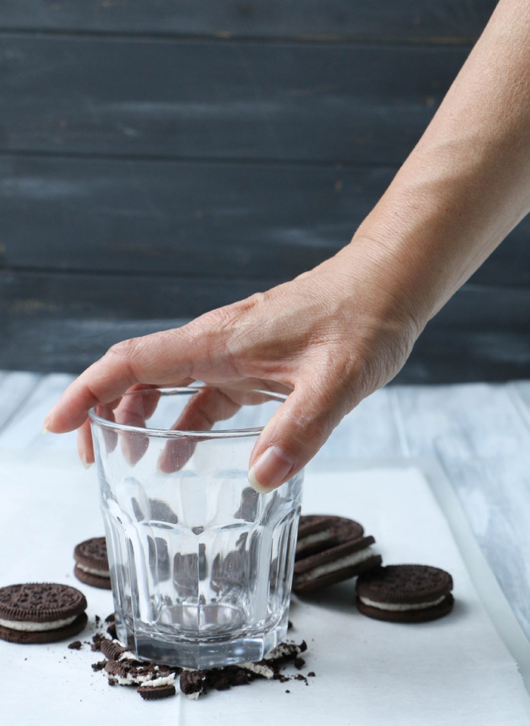 A hand crushing peppermint sandwich cookies with a drinking glass.