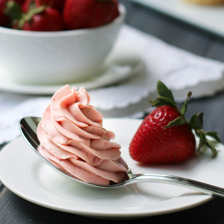 Close up of strawberry Whipped Cream Cream Cheese Frosting piped in a swirl on a spoon that's sitting on a white plate with a fresh strawberry