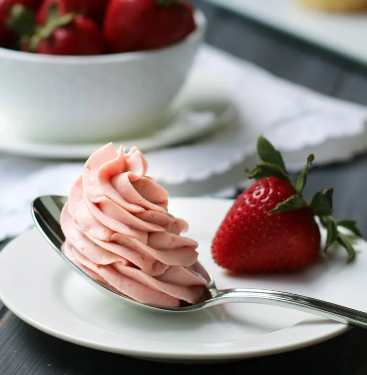 Close up of strawberry Whipped Cream Cream Cheese Frosting piped in a swirl on a spoon that's sitting on a white plate with a fresh strawberry