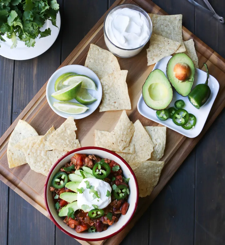 A overhead photo of a bowl of Turkey Chili topped with slices of avocado, jalapeno, sour cream and cilantro surrounded by nachos chips.